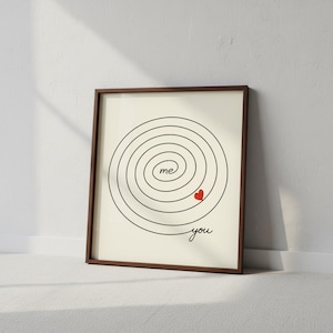 Op de afbeelding: Ingelijste kunstdruk met een minimalistisch ontwerp. Een zwarte spiraal omcirkelt het woord "me" en een rood hart, met het woord "you" onderaan. De lijst is bruin en de achtergrond is gebroken wit.