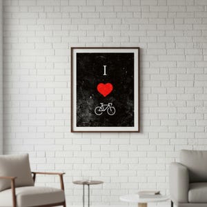 Op de afbeelding: Ingelijste kunstdruk met een zwarte achtergrond met de woorden "I ❤️" boven een witte fietsillustratie. Het hart is rood. De lijst is bruin. De print wordt weergegeven op een witte bakstenen muur.