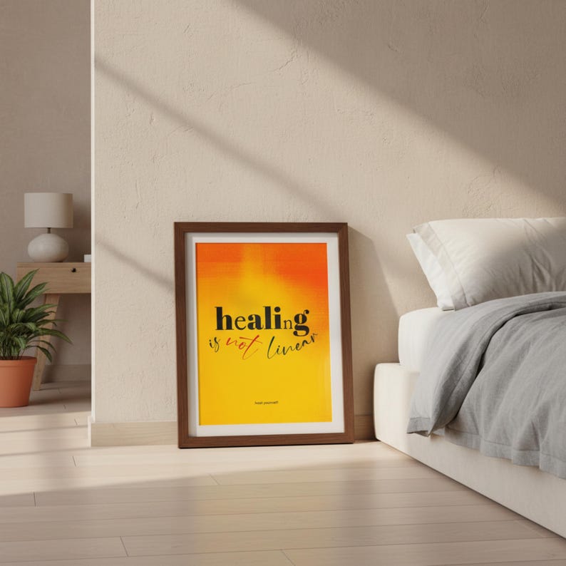 May include: A framed print with the words "healing is not linear" in black script on a gradient orange and yellow background. The print is in a brown frame and leans against a wall near a bed.