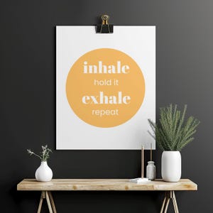 Peut inclure: Une affiche blanche avec les mots "inhale, hold it, exhale, repeat" en texte blanc sur un cercle jaune, maintenue par une pince noire. L'affiche est au-dessus d'une table en bois avec des vases blancs et de la verdure.
