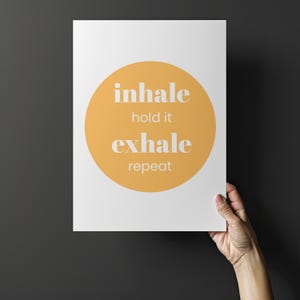 Peut inclure: Une affiche blanche avec un grand cercle orange au centre. Les mots "inhale, hold it, exhale, repeat" sont imprim&eacute;s en blanc, centr&eacute;s dans le cercle. L'affiche est tenue par une main sur un fond gris fonc&eacute;.
