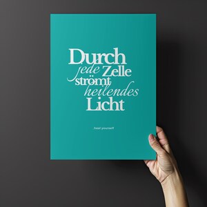 May include: A teal poster with white text that reads "Durch jede Zelle str&ouml;mt heilendes Licht" and "heal yourself" in a smaller font. The poster is held by a hand against a dark grey background.