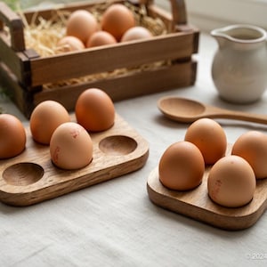 May include: A wooden crate filled with brown eggs and straw. Two wooden egg holders, each holding four eggs, are in the foreground. A wooden spoon and a small white jug are also present, suggesting a kitchen or breakfast setting.