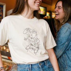 May include: Cream-colored t-shirt featuring a black line drawing of a video game controller design. The shirt is paired with blue jeans. Two people are smiling and laughing in the image.
