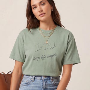 May include: Sage green t-shirt with the text "1+1=2 keep life simple" in black script. The model is wearing light wash blue jeans and gold jewellery. A brown leather tote bag is visible in the background.