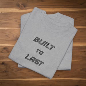 May include: Two light gray t-shirts are neatly folded on a wooden surface. The top shirt displays the text "BUILT TO LAST" in a distressed, black font. The shirts are made of a soft material and are ready to wear.