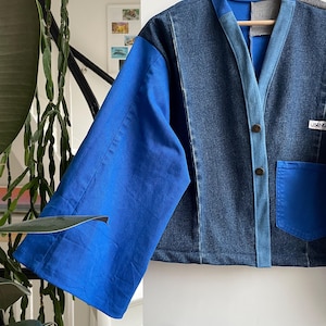 以下が含まれることがあります： 青とデニムのパッチワークジャケット。鮮やかな青色の袖と、ポケットが1つ付いたデニムのボディが特徴です。木製のハンガーに掛けられています。「USENG」と書かれたラベルが付いています。