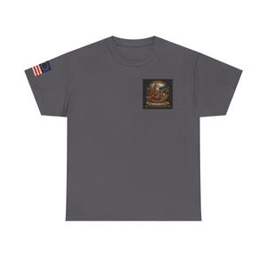 Op de afbeelding: Een donkergrijs t-shirt met een kleine vierkante patch met een gedetailleerde illustratie van een geweer, een persoon en tekst. De linkermouw toont een kleine Amerikaanse vlag. Het shirt is gemaakt van een zacht materiaal.