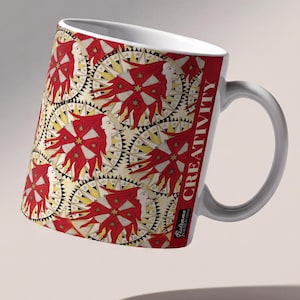 May include: A white ceramic mug featuring a repeating pattern of stylized faces with flowing red hair and golden star accents set against circular sunburst motifs. The word CREATIVITY is printed vertically in white on a red background panel.