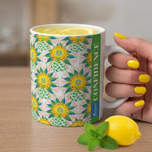 May include: A white ceramic mug featuring a repeating pattern of yellow, blue, and green floral-like mandalas with coral-colored accents. A vertical green stripe on the side contains the white text CONFIDENCE and a small blue logo below it.
