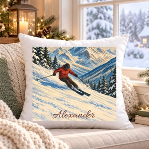Könnte beinhalten: Ein weißes quadratisches Kissen mit einer gedruckten Illustration eines Skifahrers in roter Jacke und schwarzer Hose, der einen verschneiten Berghang mit Tannen hinunterfährt. Der Name Alexander ist unten in brauner Schreibschrift gedruckt.