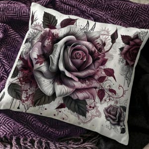 Può includere: Un cuscino decorativo con un design a rosa dettagliato nei toni del bianco, grigio e bordeaux. Il motivo floreale comprende foglie e illustrazioni di rose più piccole su sfondo bianco. Il cuscino è posto su una coperta viola.