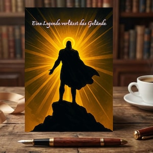 Peut inclure: Carte de vœux avec une silhouette d'une figure avec une cape, debout sur un rocher, sur un fond jaune rayonnant. Le texte allemand "Eine Legende verlässt das Gelände" est en haut. La carte est sur une surface en bois avec un stylo et une tasse de café.