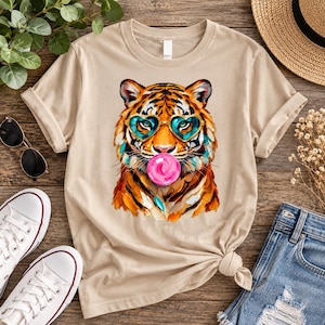 Könnte beinhalten: Ein beiges T-Shirt mit Rundhalsausschnitt und einem Grafikdruck eines Tigers, der eine türkisfarbene herzförmige Brille trägt und eine leuchtend rosa Kaugummiblase bläst. Die Tigerillustration verwendet lebendige Orange-, Schwarz- und Türkistöne.