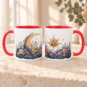 May include: Two white ceramic mugs with bright red handles and interiors. The left mug features a golden crescent moon surrounded by colorful flowers. The right mug displays a golden starburst design above a cluster of purple, pink, and blue flowers.