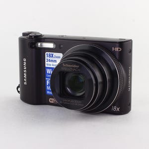 Puede incluir: C&aacute;mara digital Samsung negra con una lente prominente y un flash incorporado. La c&aacute;mara tiene un objetivo zoom y muestra el texto "18x Zoom 24mm Wide Angle". La c&aacute;mara est&aacute; sobre un fondo blanco.