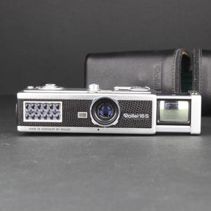 Puede incluir: Una cámara Rollei 16S vintage con cuerpo, lente y visor plateados y negros. La cámara va acompañada de una funda de cuero negra. El texto "MADE IN GERMANY BY ROLLEI" es visible en la cámara.