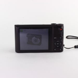 Puede incluir: Una c&aacute;mara digital negra con una pantalla que muestra una imagen de una c&aacute;mara. La c&aacute;mara tiene un bot&oacute;n rojo y botones de control. Una correa de mu&ntilde;eca negra est&aacute; unida al lateral.