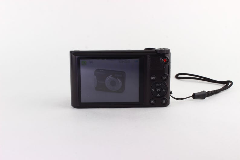 Puede incluir: Una c&aacute;mara digital negra con una pantalla que muestra una imagen de una c&aacute;mara. La c&aacute;mara tiene un bot&oacute;n rojo y botones de control. Una correa de mu&ntilde;eca negra est&aacute; unida al lateral.