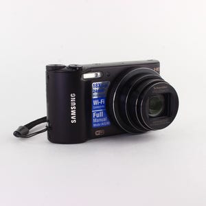 Puede incluir: C&aacute;mara digital Samsung negra con objetivo retr&aacute;ctil y correa para la mu&ntilde;eca. La c&aacute;mara cuenta con zoom 18x, Wi-Fi y modo manual completo. El logotipo de Samsung es visible en el lateral.