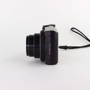 Puede incluir: Una c&aacute;mara digital negra con una lente prominente y una correa de mu&ntilde;eca negra. El cuerpo de la c&aacute;mara tiene el texto "14.2 MEGA PIXELS" impreso. La c&aacute;mara est&aacute; colocada sobre una superficie blanca.