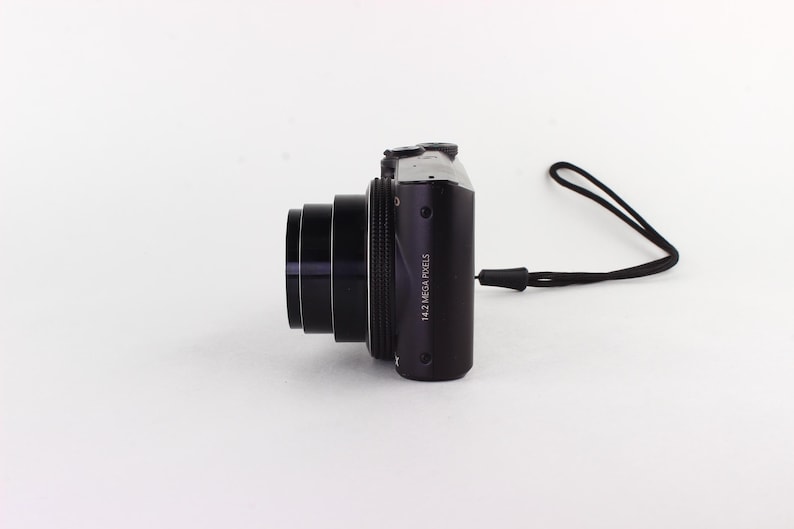 Puede incluir: Una c&aacute;mara digital negra con una lente prominente y una correa de mu&ntilde;eca negra. El cuerpo de la c&aacute;mara tiene el texto "14.2 MEGA PIXELS" impreso. La c&aacute;mara est&aacute; colocada sobre una superficie blanca.
