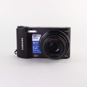 Puede incluir: Una c&aacute;mara digital Samsung negra con una lente prominente y el texto "18X Zoom" y "Wi-Fi". La c&aacute;mara tiene un flash incorporado y una lente Schneider. La c&aacute;mara est&aacute; sobre un fondo blanco.