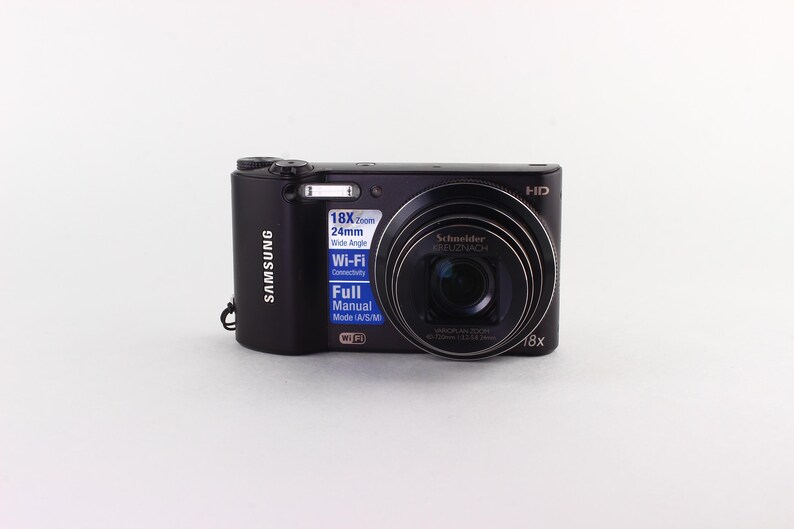 Puede incluir: Una c&aacute;mara digital Samsung negra con una lente prominente y el texto "18X Zoom" y "Wi-Fi". La c&aacute;mara tiene un flash incorporado y una lente Schneider. La c&aacute;mara est&aacute; sobre un fondo blanco.