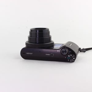 Puede incluir: C&aacute;mara digital negra con objetivo retr&aacute;ctil y correa de mu&ntilde;eca negra. La c&aacute;mara tiene un bot&oacute;n de encendido y varios diales de control. El n&uacute;mero de modelo WB150F es visible en el cuerpo.