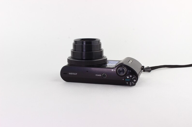 Puede incluir: C&aacute;mara digital negra con objetivo retr&aacute;ctil y correa de mu&ntilde;eca negra. La c&aacute;mara tiene un bot&oacute;n de encendido y varios diales de control. El n&uacute;mero de modelo WB150F es visible en el cuerpo.