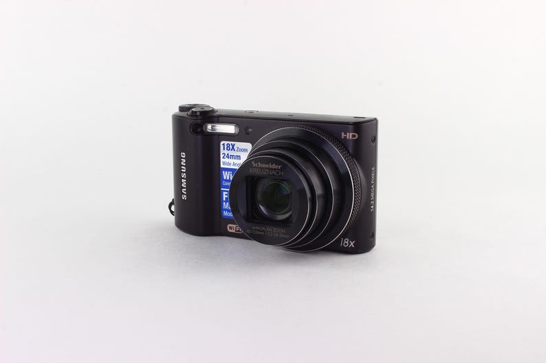 Puede incluir: C&aacute;mara digital Samsung negra con una lente prominente y un flash incorporado. La c&aacute;mara tiene un objetivo zoom y muestra el texto "18x Zoom 24mm Wide Angle". La c&aacute;mara est&aacute; sobre un fondo blanco.