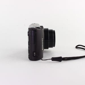 Puede incluir: C&aacute;mara digital Samsung negra con lente y correa de mu&ntilde;eca negra. La c&aacute;mara est&aacute; colocada horizontalmente, mostrando su perfil lateral sobre un fondo blanco. El logotipo de Samsung es visible.