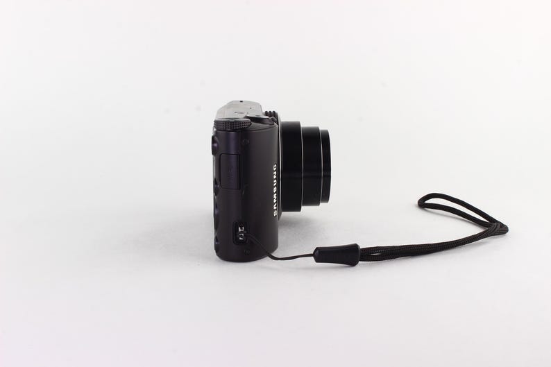 Puede incluir: C&aacute;mara digital Samsung negra con lente y correa de mu&ntilde;eca negra. La c&aacute;mara est&aacute; colocada horizontalmente, mostrando su perfil lateral sobre un fondo blanco. El logotipo de Samsung es visible.