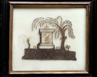 May include: A framed silhouette of a tombstone with a cross on top, surrounded by a weeping willow tree and other foliage, all made from human hair.