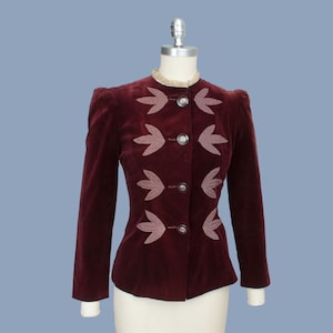 May include: A burgundy velvet jacket with leaf-shaped appliqués and silver buttons. The jacket has a high collar and long sleeves. The garment is displayed on a mannequin against a light blue background.