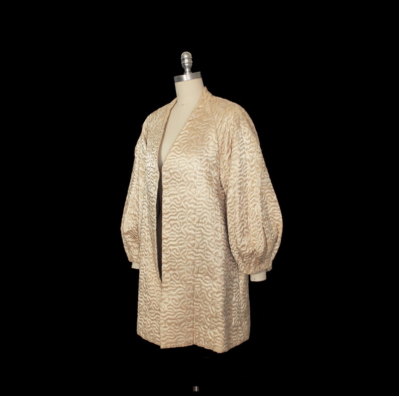 May include: A long, cream-colored quilted jacket with a V-neck and long sleeves. The jacket has a textured, wavy pattern.