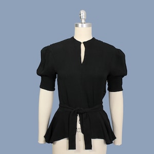 May include: A black, short-sleeved blouse with a keyhole neckline and a fitted bodice. The blouse has a wide, gathered peplum hem and a black fabric belt.