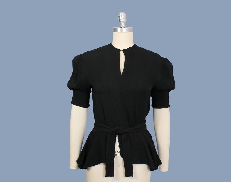 May include: A black, short-sleeved blouse with a keyhole neckline and a fitted bodice. The blouse has a wide, gathered peplum hem and a black fabric belt.