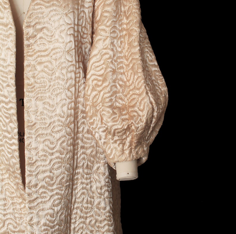 May include: A long, cream-colored quilted robe with a shawl collar and long sleeves. The robe has a unique, swirling pattern.