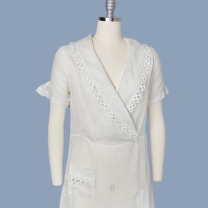 May include: A white dress with a wrap-style neckline and short sleeves. The dress features delicate lace trim along the collar and pocket, and a subtle dotted pattern. The dress is displayed on a mannequin.
