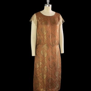 May include: A vintage brown dress with a gold diamond pattern. The dress has short sleeves with lace trim and a round neckline. The dress is displayed on a mannequin against a black background.