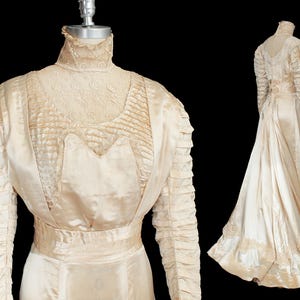 Peut inclure: Robe de mariée vintage crème avec un col montant et des manches longues. La robe présente des détails de dentelle complexes sur le corsage et les manches, avec une jupe et une traîne fluides. La robe est présentée sur un mannequin sur fond noir.