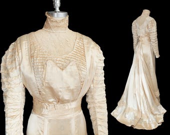 May include: A cream-coloured vintage wedding dress with a high neckline and long sleeves. The dress features intricate lace detailing on the bodice and sleeves, with a flowing skirt and train. The dress is displayed on a mannequin against a black background.