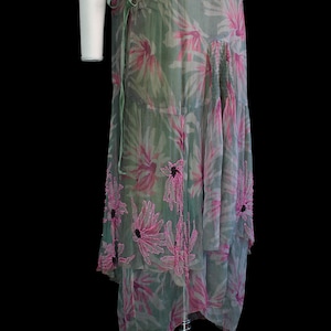 May include: A flowing, sheer dress in shades of green and pink, adorned with large floral patterns. The dress features a tiered skirt and a delicate green tie at the waist. The floral details are accented with darker centers.