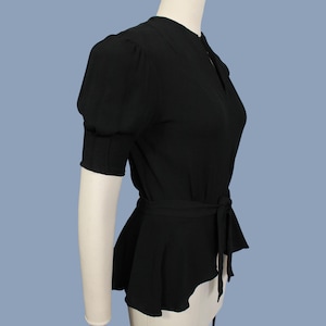 May include: A black, short-sleeved blouse with a cinched waist and a peplum hem. The blouse has a high neckline and a button closure at the front.