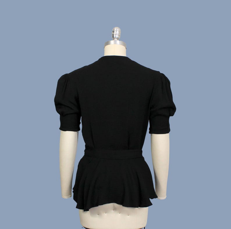 May include: A black, short-sleeved blouse with a fitted bodice and a gathered, flowing skirt. The blouse has a V-neckline and a belt at the waist.