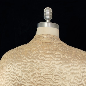 May include: A cream-colored quilted jacket with a detailed pattern of stitching. The jacket is on a dress form with a silver head.