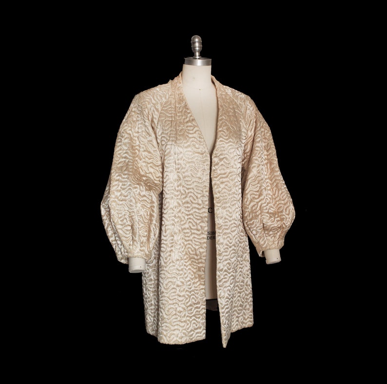 May include: A cream-colored quilted jacket with long sleeves and a V-neck. The jacket has a textured, wavy pattern and a slightly puffed sleeve.