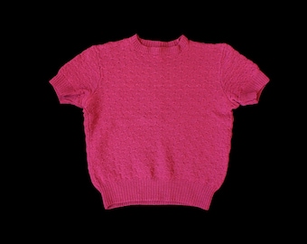 May include: A short-sleeved, magenta-colored knit top. The top features a textured pattern and a ribbed hem and neckline. The garment is displayed against a black background, highlighting its color and texture. The top is a casual wear item.
