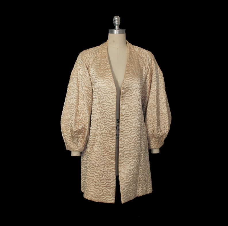 May include: A long, cream-colored jacket with a quilted pattern and large, puffed sleeves. The jacket has a shawl collar and a front closure.